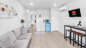 Un salon moderne avec un canapé gris, un coin repas avec une table et des chaises en bois, une cuisine avec un réfrigérateur bleu et un téléviseur fixé au mur qui affiche le logo « HCPL ».