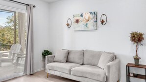 Un salon moderne avec un canapé de couleur claire, une porte en verre donnant sur un espace extérieur, un tableau accroché au mur et une petite table sur laquelle est posée une plante.