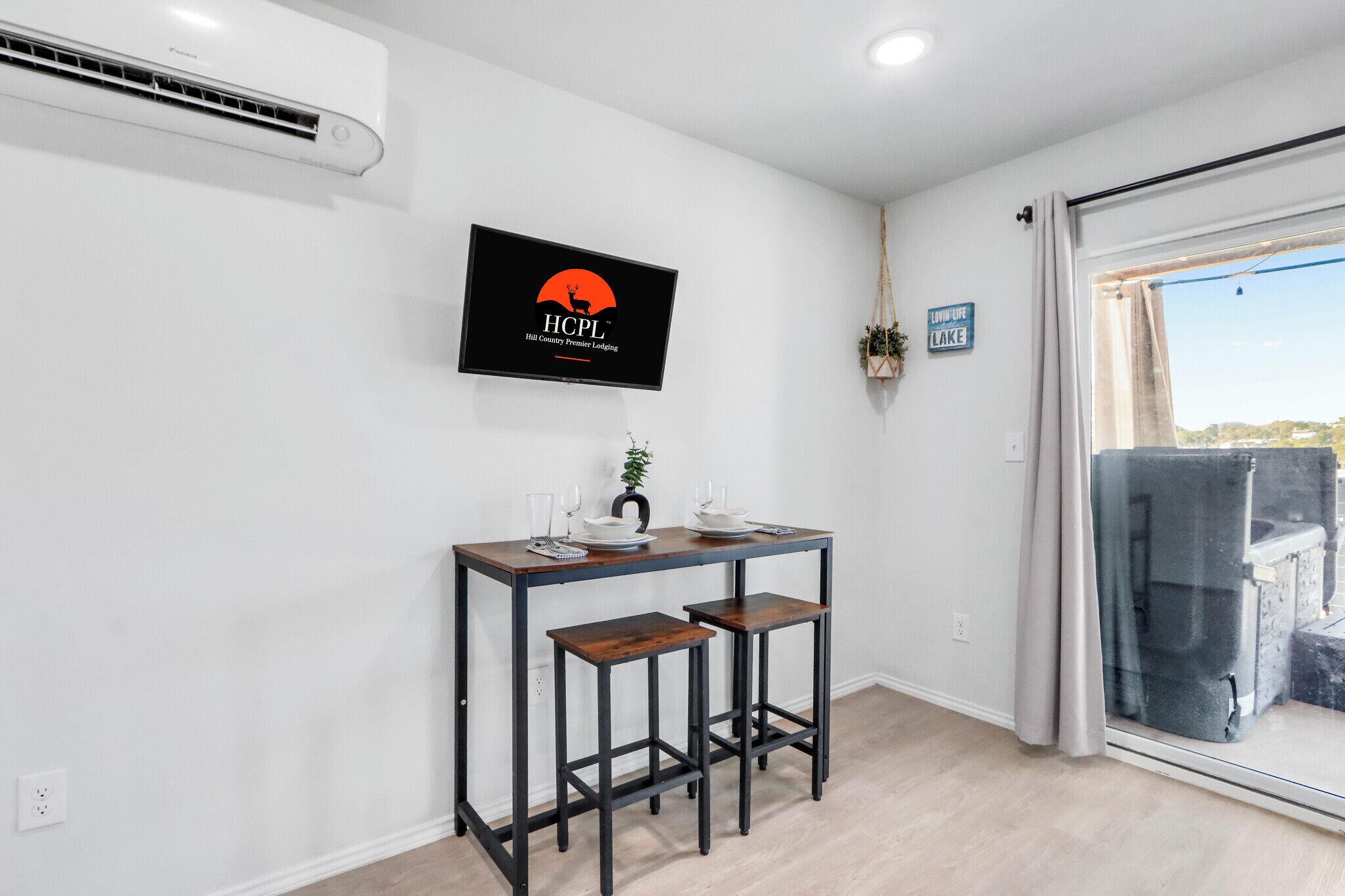 Une pièce moderne comprenant une table à manger, deux tabourets de bar, un téléviseur fixé au mur qui affiche un logo, et une vue sur un lac, visible à travers une porte-fenêtre coulissante.