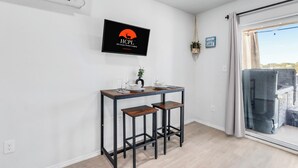 Une pièce moderne comprenant une table à manger, deux tabourets de bar, un téléviseur fixé au mur qui affiche un logo, et une vue sur un lac, visible à travers une porte-fenêtre coulissante.