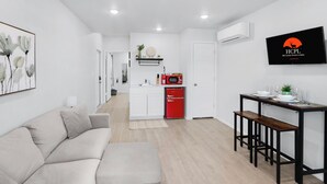 Una sala moderna con un sofá, una mesa de comedor con bancos, una zona de cocina con un refrigerador rojo y un televisor montado que muestra 'HCPL'.