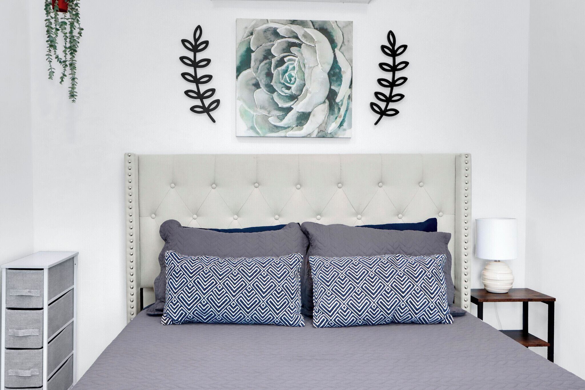 A bedroom with a tufted headboard, two patterned pillows, a wall-mounted artwork, and a bedside table with a lamp.