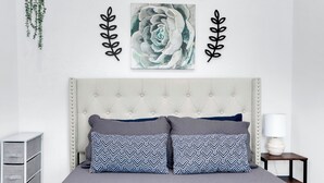 Un dormitorio con cabecera capitonada, dos almohadas estampadas, una obra de arte colgada en la pared y una mesita de noche con lámpara.