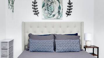 A bedroom with a tufted headboard, two patterned pillows, a wall-mounted artwork, and a bedside table with a lamp.