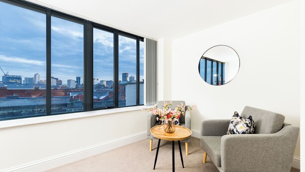 Ein modernes Wohnzimmer mit einem großen Fenster, einem grauen Sessel, einem runden Couchtisch mit Blumenvase und einem runden Spiegel an der Wand.
