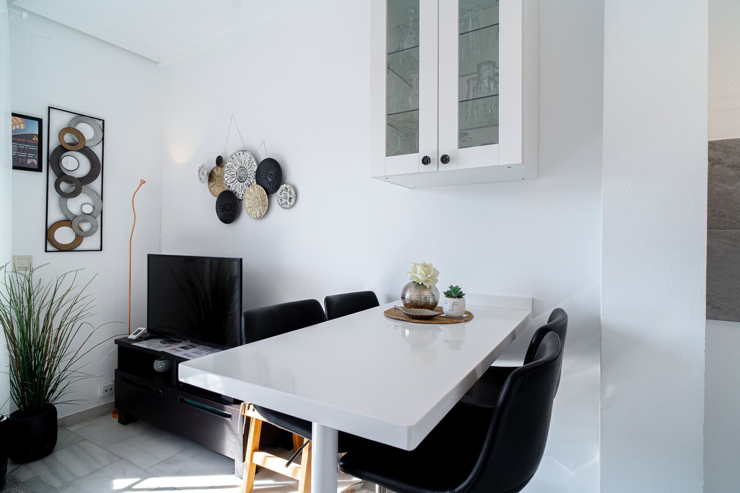 Zona pranzo moderna con tavolo bianco, sedie nere, televisore e decorazioni a parete.