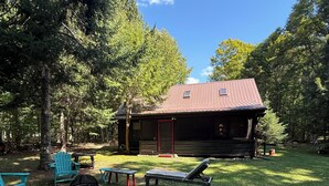 A cabin with a red door, surrounded by a grassy yard, a fire pit, and outdoor furniture. - Hoel Pond Hideaway@Saranac Inn (Saranac Lake)
