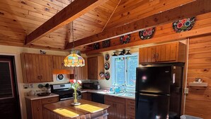 A kitchen with wooden cabinets, a black refrigerator, a wooden island with stools, a hanging light fixture, and a window with a view. - Hoel Pond Hideaway@Saranac Inn (Saranac Lake)