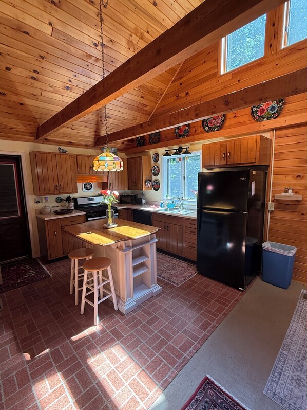 Una cocina con armarios de madera, un refrigerador negro, una isla de madera con taburetes, una lámpara colgante y una ventana con vistas.