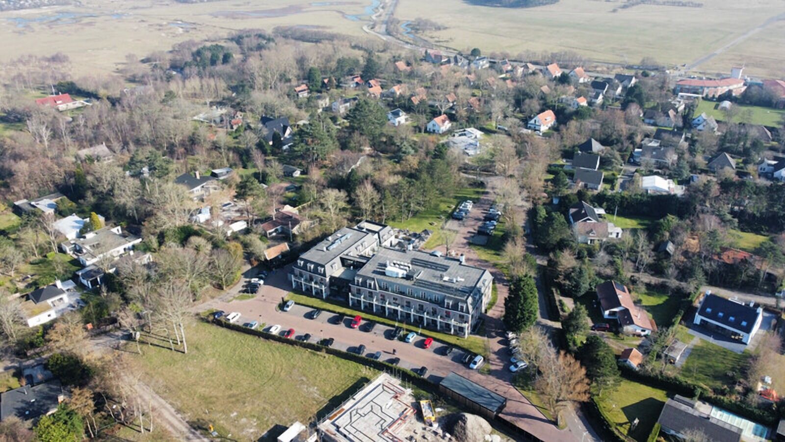 An aerial view of a residential area with a large building that appears to be a commercial or institutional structure, surrounded by houses and greenery.
