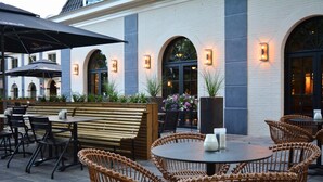 Een terras met rieten stoelen en tafels, een houten bank en potplanten.