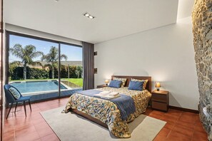 Quarto com cama grande, mesa de cabeceira, cadeira e vista para uma piscina.
