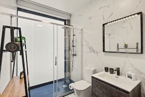 Une salle de bain moderne avec une douche à parois de verre, un mur en marbre, un grand miroir et une étagère en bois ornée d’une plante.
