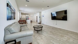 A modern living room with a sofa, a coffee table, a flat-screen TV, and a kitchen in the background.