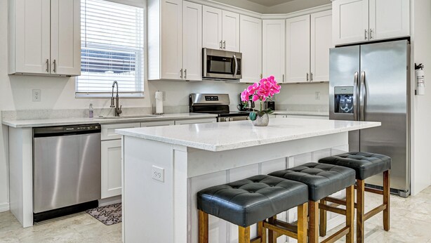 A modern kitchen with white cabinets, stainless steel appliances, a central island with stools, and a window with blinds.