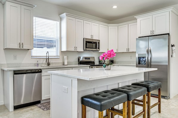 A modern kitchen with white cabinets, stainless steel appliances, a central island with stools, and a window with blinds. - Brand New! Modern Tampa Getaway w/ Private Yard (Tampa)