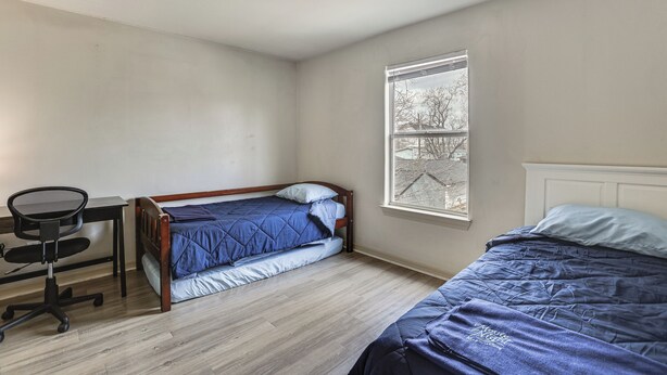 Eine kleine Einzimmerwohnung mit einem Holztisch, einem Stuhl, einem Bett mit blauer Bettwäsche und einem Fenster mit Blick auf Bäume.