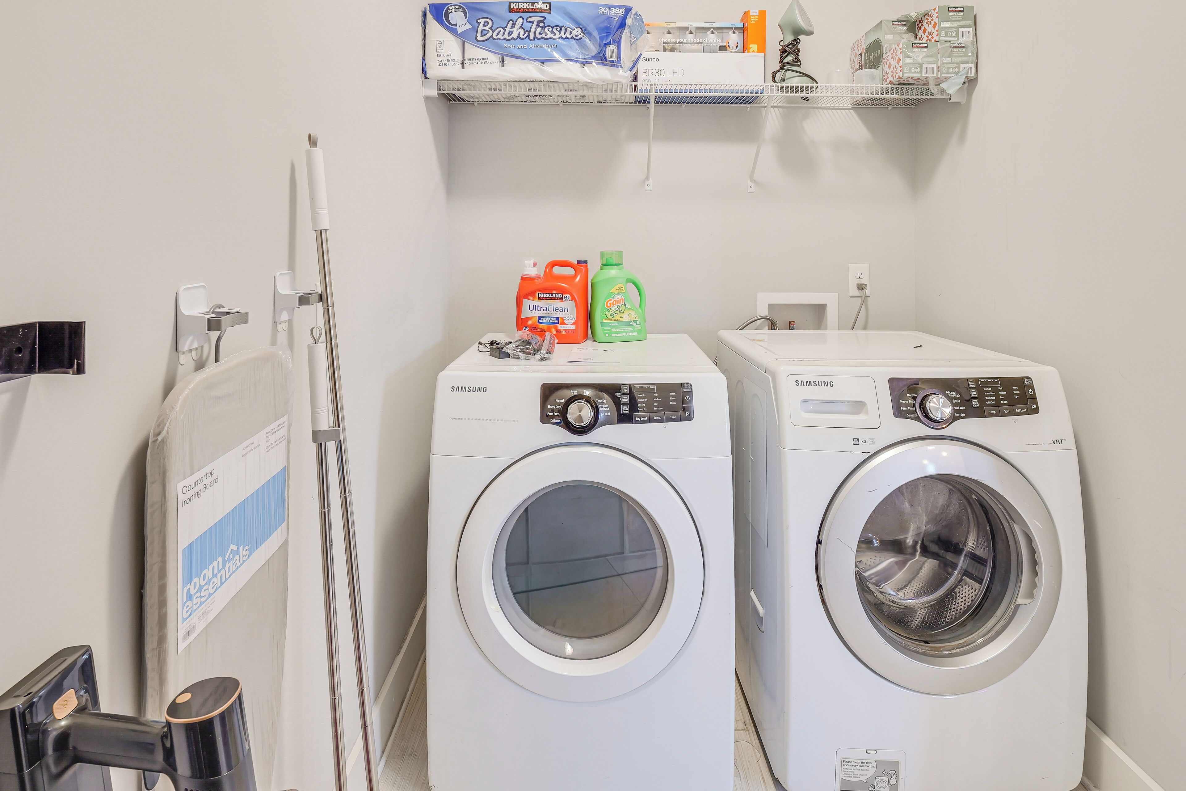 A laundry area with two front-loading washing machines, a drying rack, and a shelf with bath products.