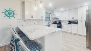A modern kitchen with white cabinets, a marble countertop, a built-in island, and a ship's wheel decoration. - 3BR Coastal Home w/ Coastal Design- Steps to Sand (Galveston)