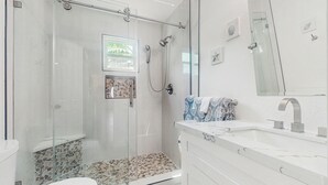 A modern bathroom with a glass-enclosed shower, a white toilet, and a marble countertop with a sink and mirror. - 3BR Coastal Home w/ Coastal Design- Steps to Sand (Galveston)