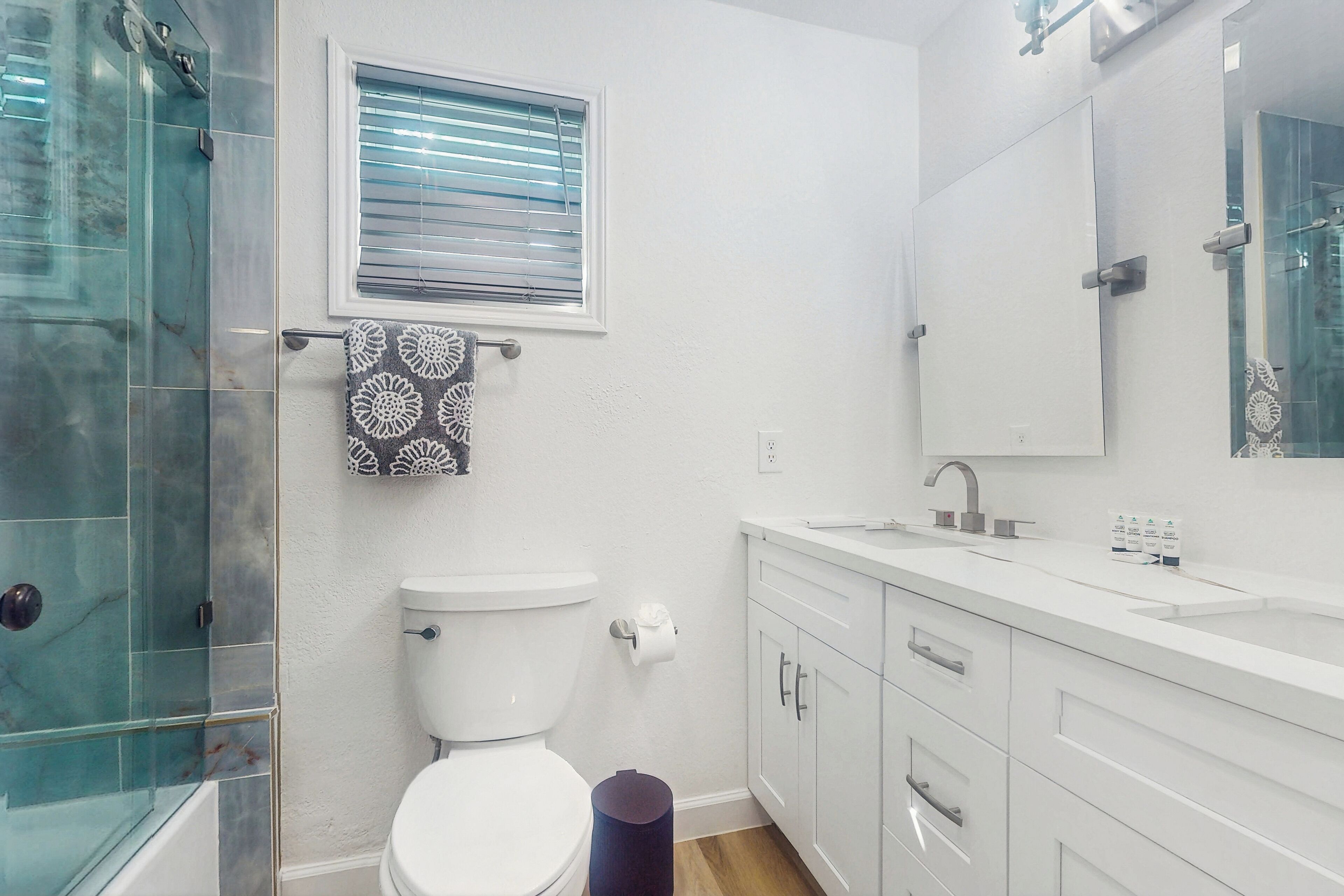 Ein Badezimmer mit Duschkabine aus Glas, Toilette, Waschbecken mit Spiegel und Fenster mit Jalousien.
