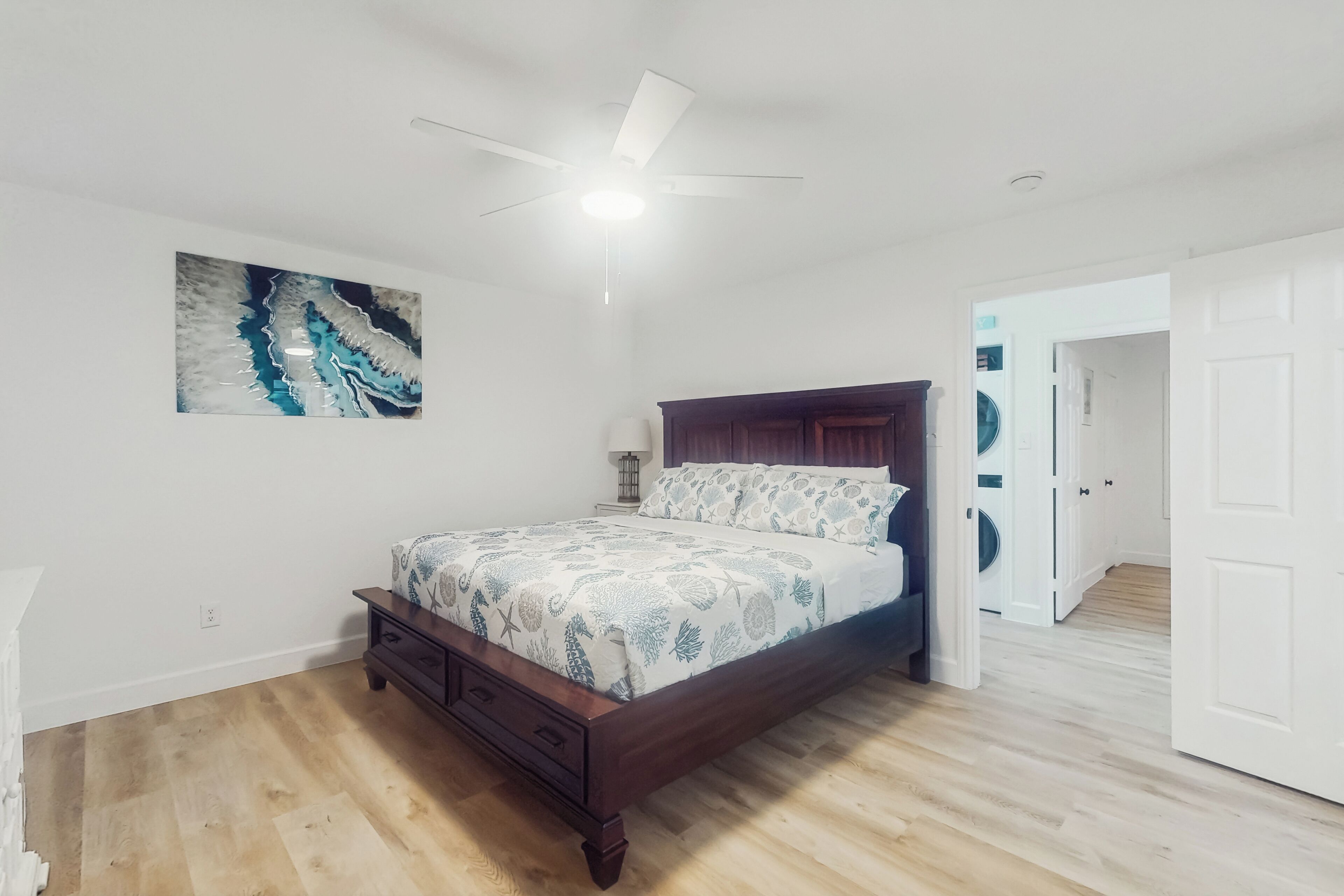 Una camera da letto con un letto di legno, una ventola a soffitto, un'opera d'arte appesa al muro e una porta che dà su un'area lavanderia.