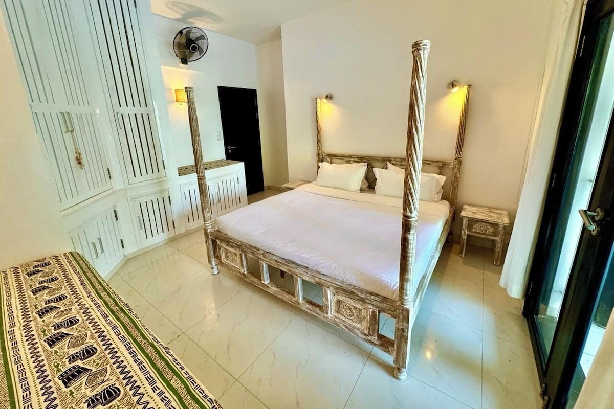 A four-poster bed with a white bedspread, a bedside table with a lamp, a radiator, and a patterned rug on the floor.