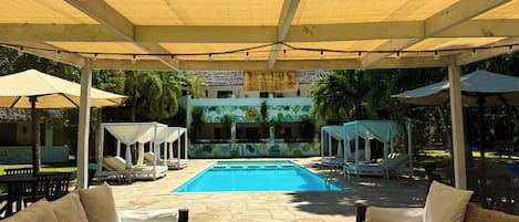 A covered outdoor pool area with lounge chairs and umbrellas, a pool, and a building with a tropical design in the background.