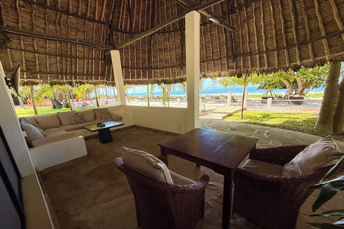 A covered outdoor seating area with a table and chairs, a sofa, and a view of the beach.