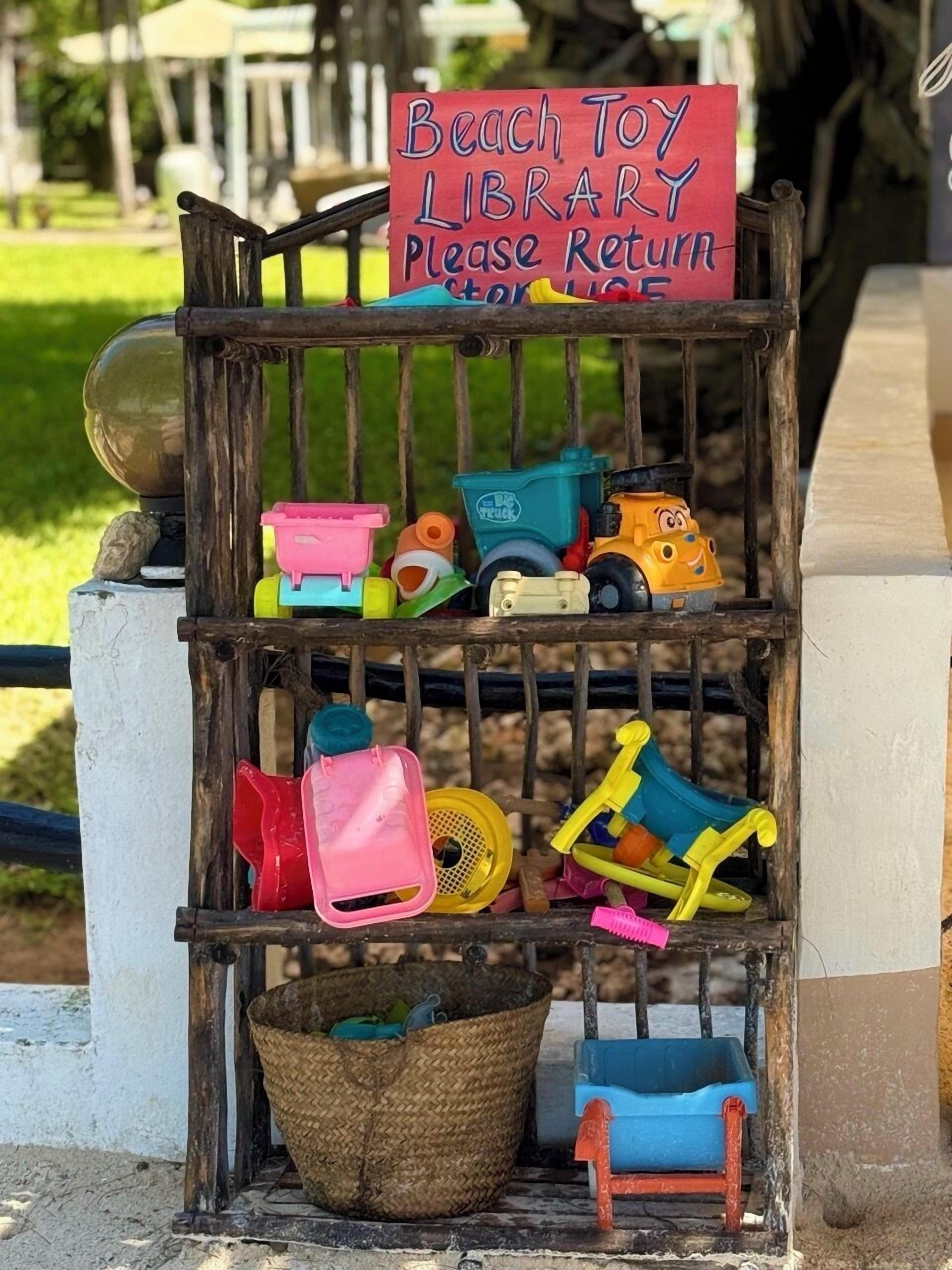 Letrero de una biblioteca de juguetes para la playa sobre un estante de madera con diversos juguetes y una cesta.
