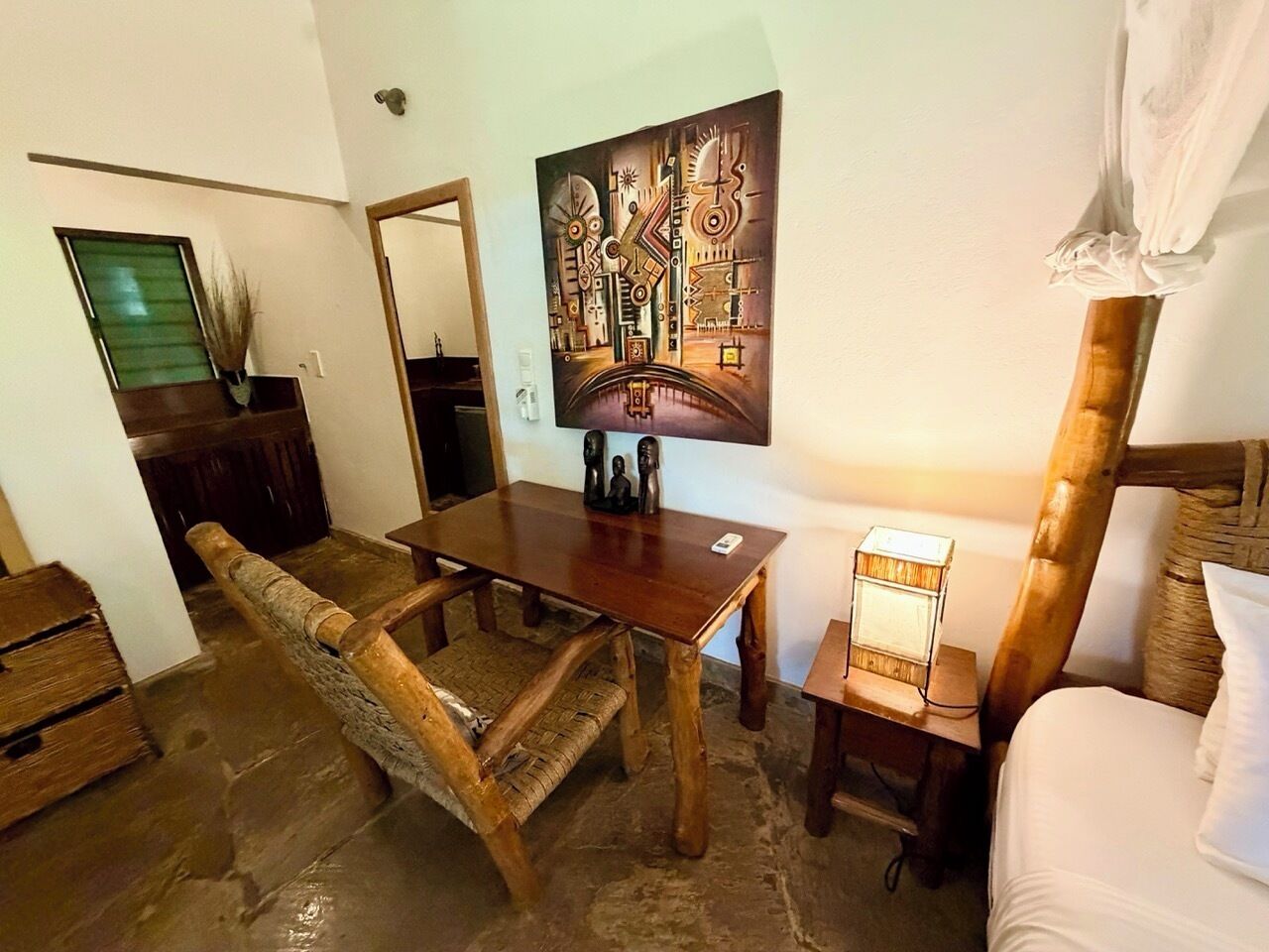 A rustic room with a wooden dining table, a woven chair, a bed with a white pillow, and a painting on the wall.