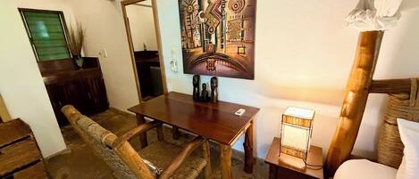 A rustic room with a wooden dining table, a woven chair, a bed with a white pillow, and a painting on the wall.