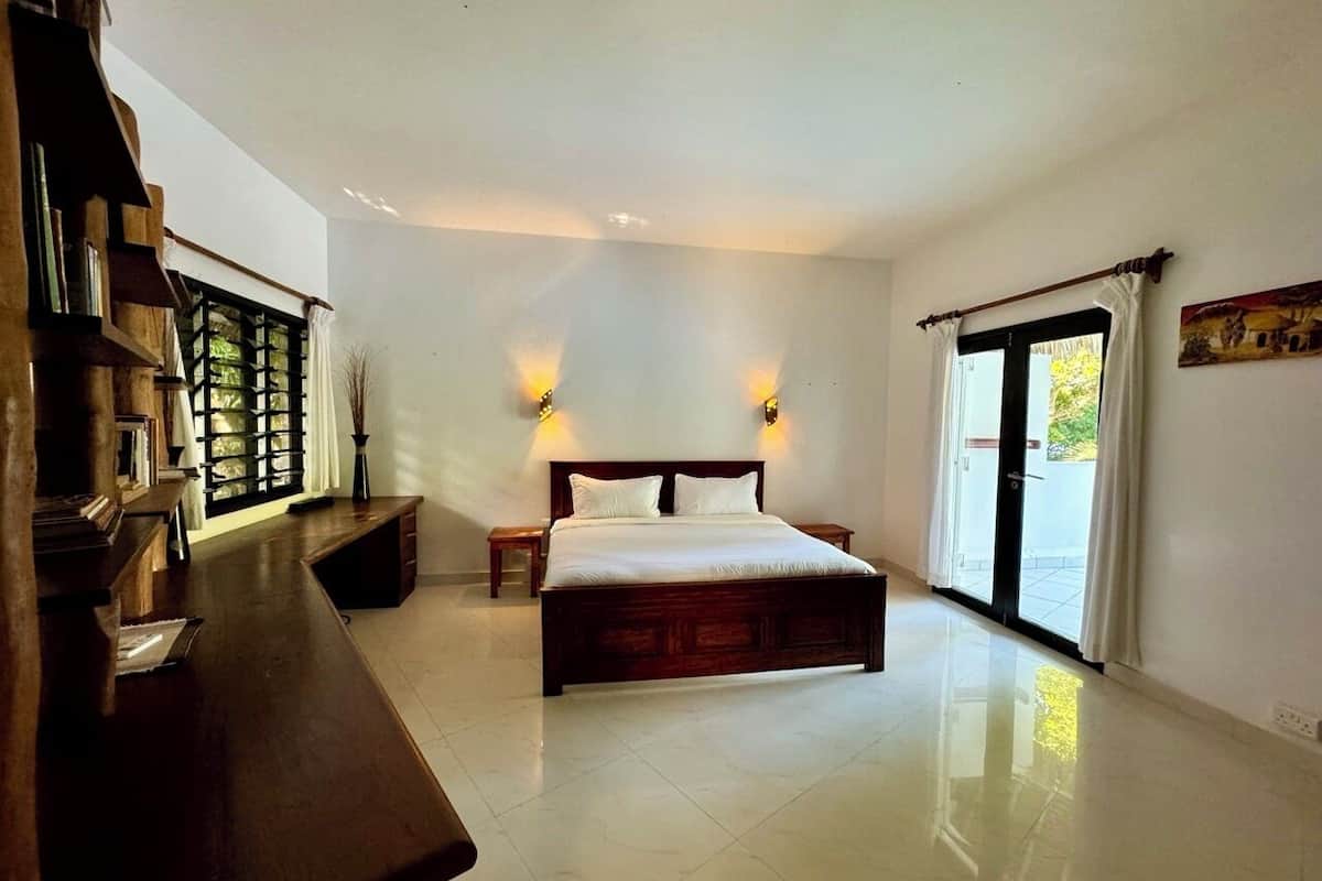 A bedroom with a large bed, a wooden desk, a window with curtains, and a door leading outside.