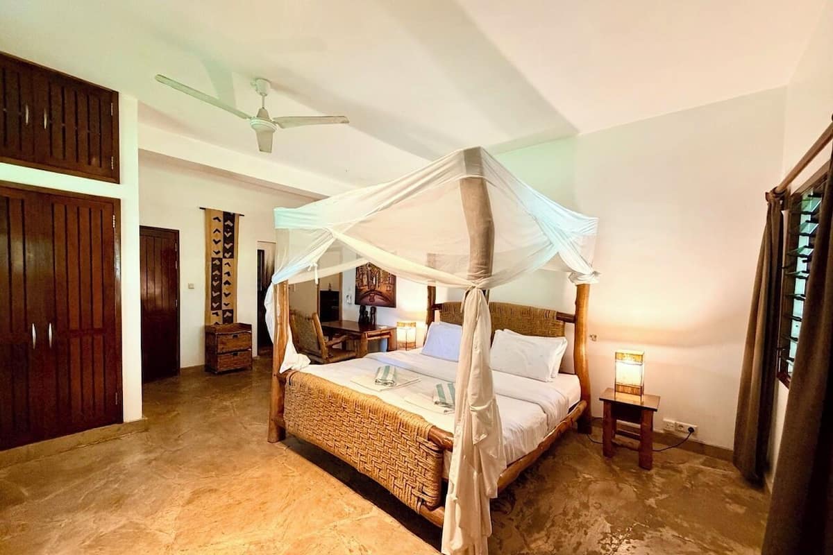 A four-poster bed with a canopy, a wooden headboard, a bedside table with a lamp, and a small wooden cabinet.