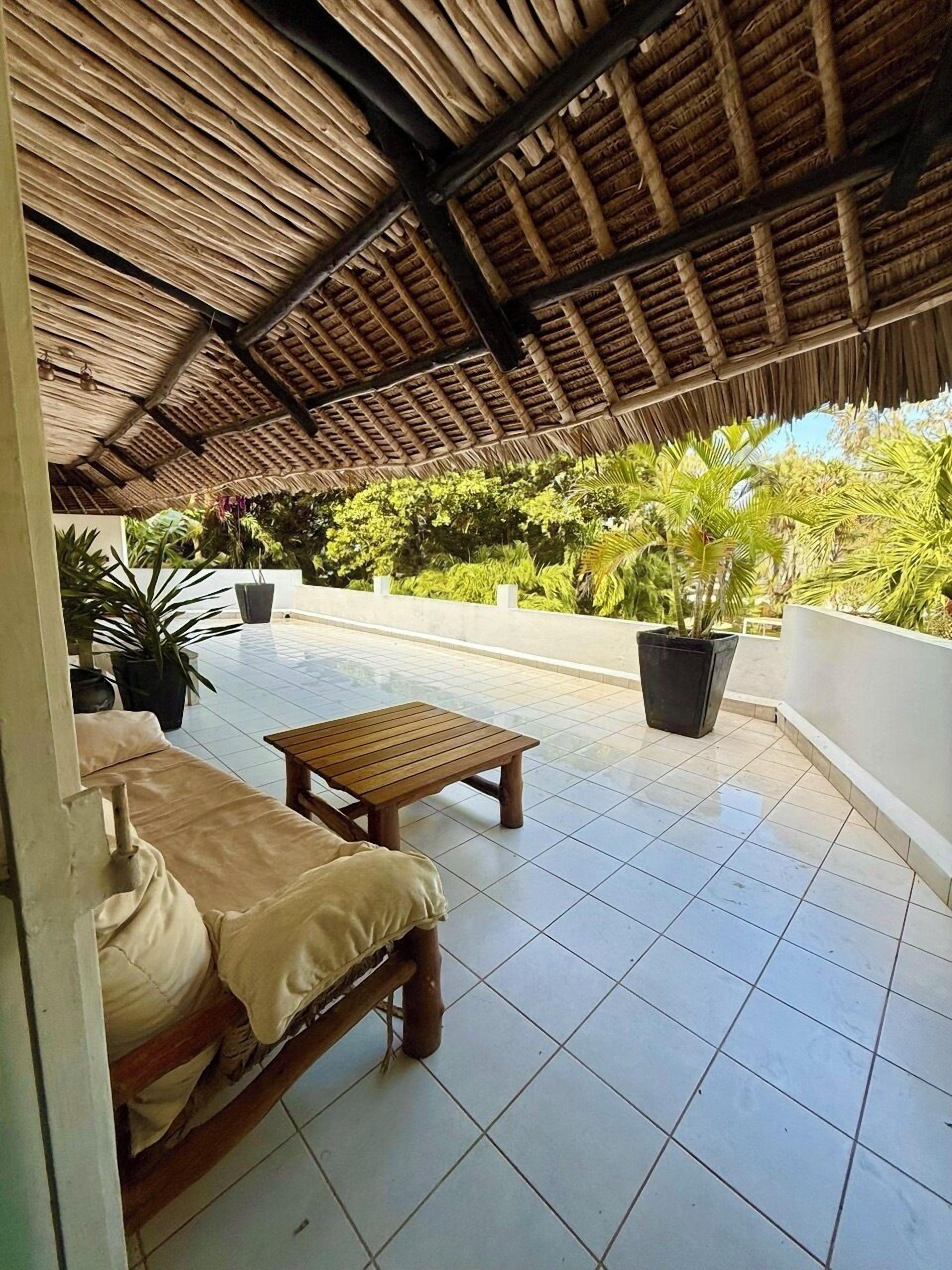 An outdoor patio with a thatched roof, tiled flooring, a wooden bench, a sofa, and potted plants.