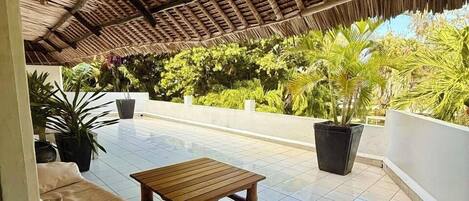 Eine Terrasse im Freien mit einem Reetdach, Fliesenboden, einer Holzbank, einer Couch und Topfpflanzen.