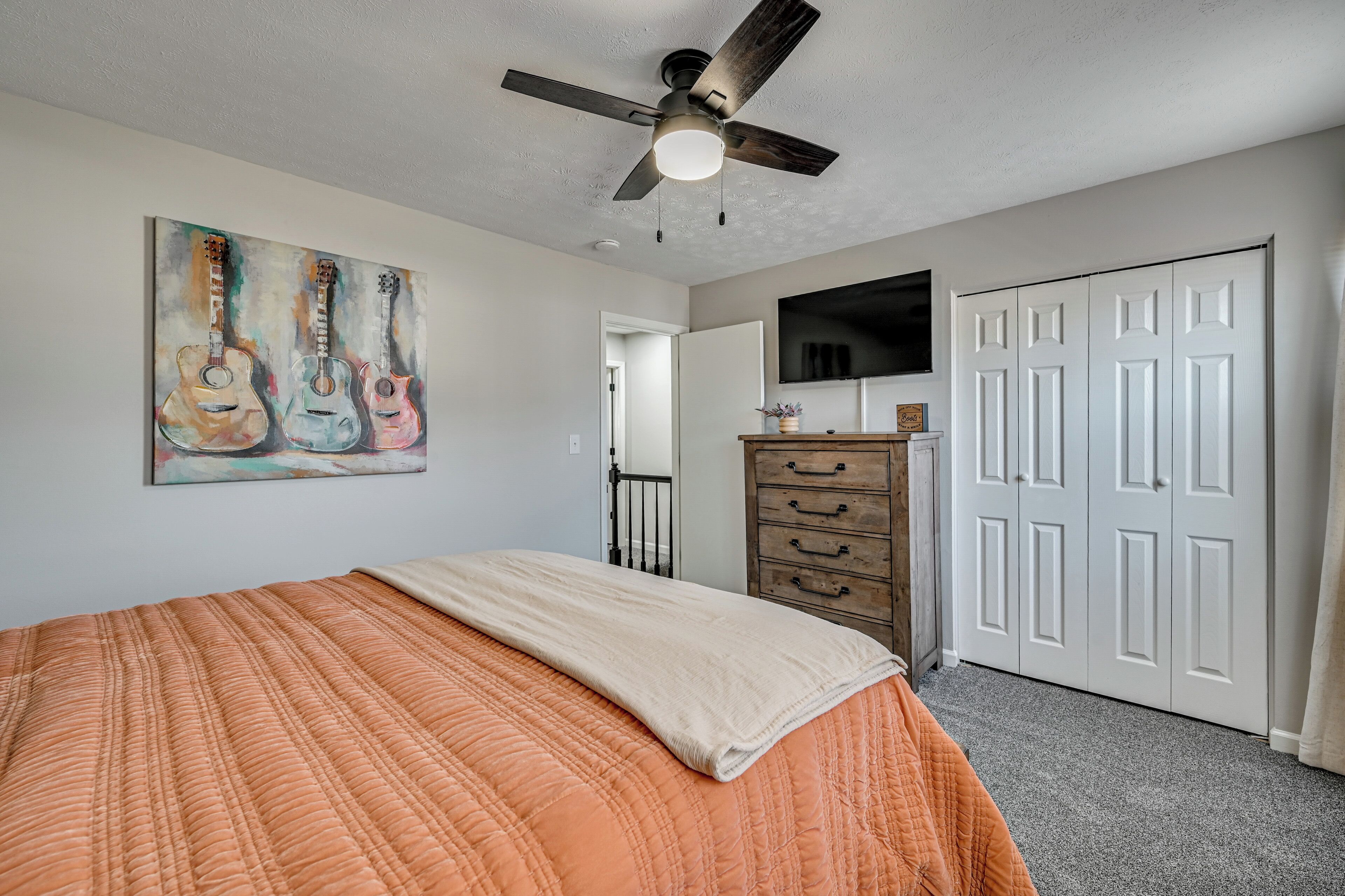 A bedroom with a bed, a ceiling fan, a painting of guitars, a dresser, and a TV on the wall.
