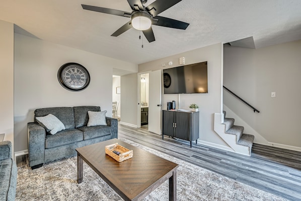 Un salon moderne avec un canapé gris, une table basse en bois, un ventilateur de plafond, une horloge, un téléviseur et un escalier.