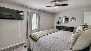 Un dormitorio con cama, ventilador de techo, televisor montado en la pared, cómoda y ventana con cortinas.