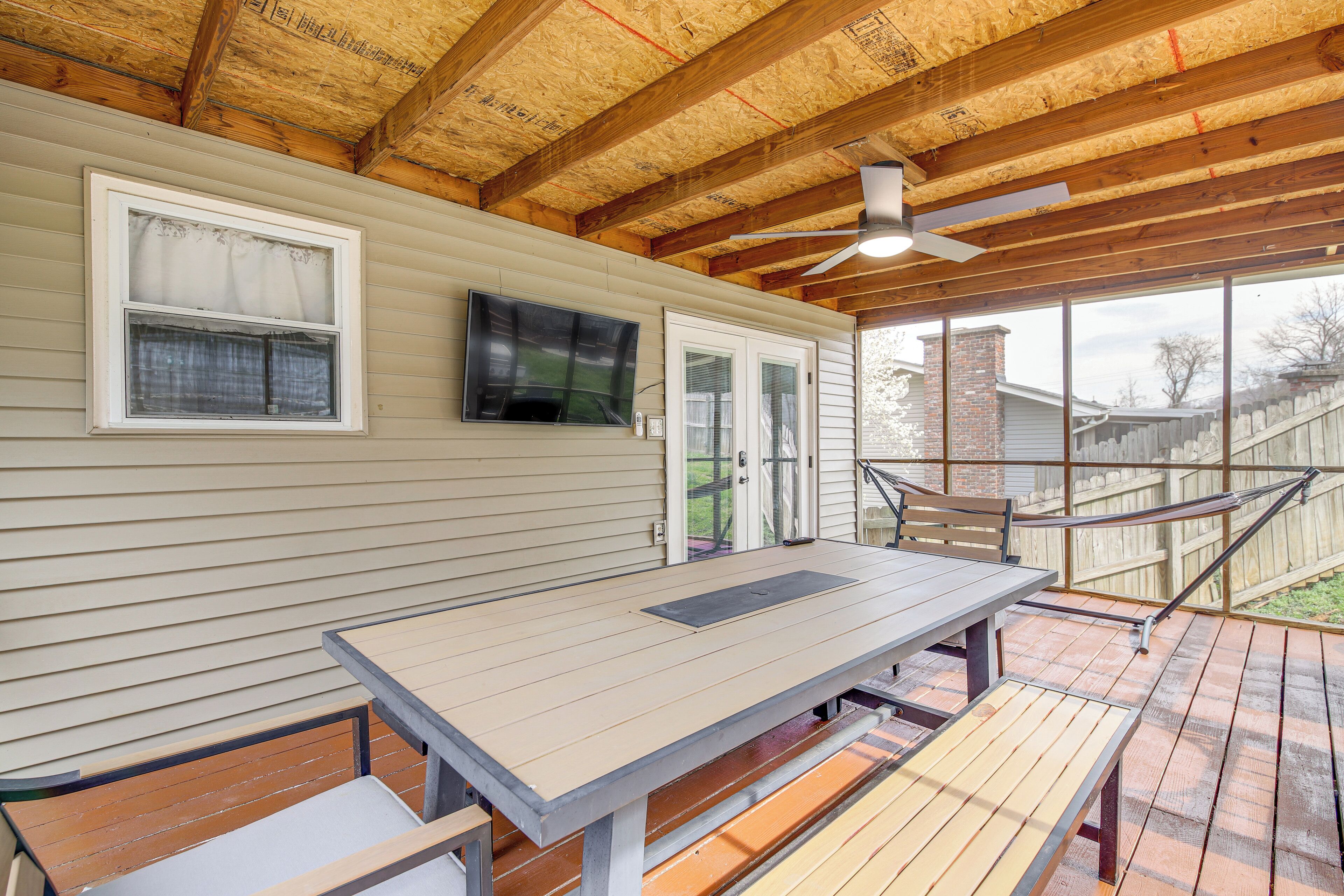 A covered patio with a picnic table and benches, wooden ceiling beams, and a TV mounted on the wall.