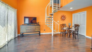 A living room with orange walls, a wooden cabinet, a TV, a dining table with chairs, and a spiral staircase. - Pool Access & Near Beach! Golf Colony Resort Condo (Surfside Beach)