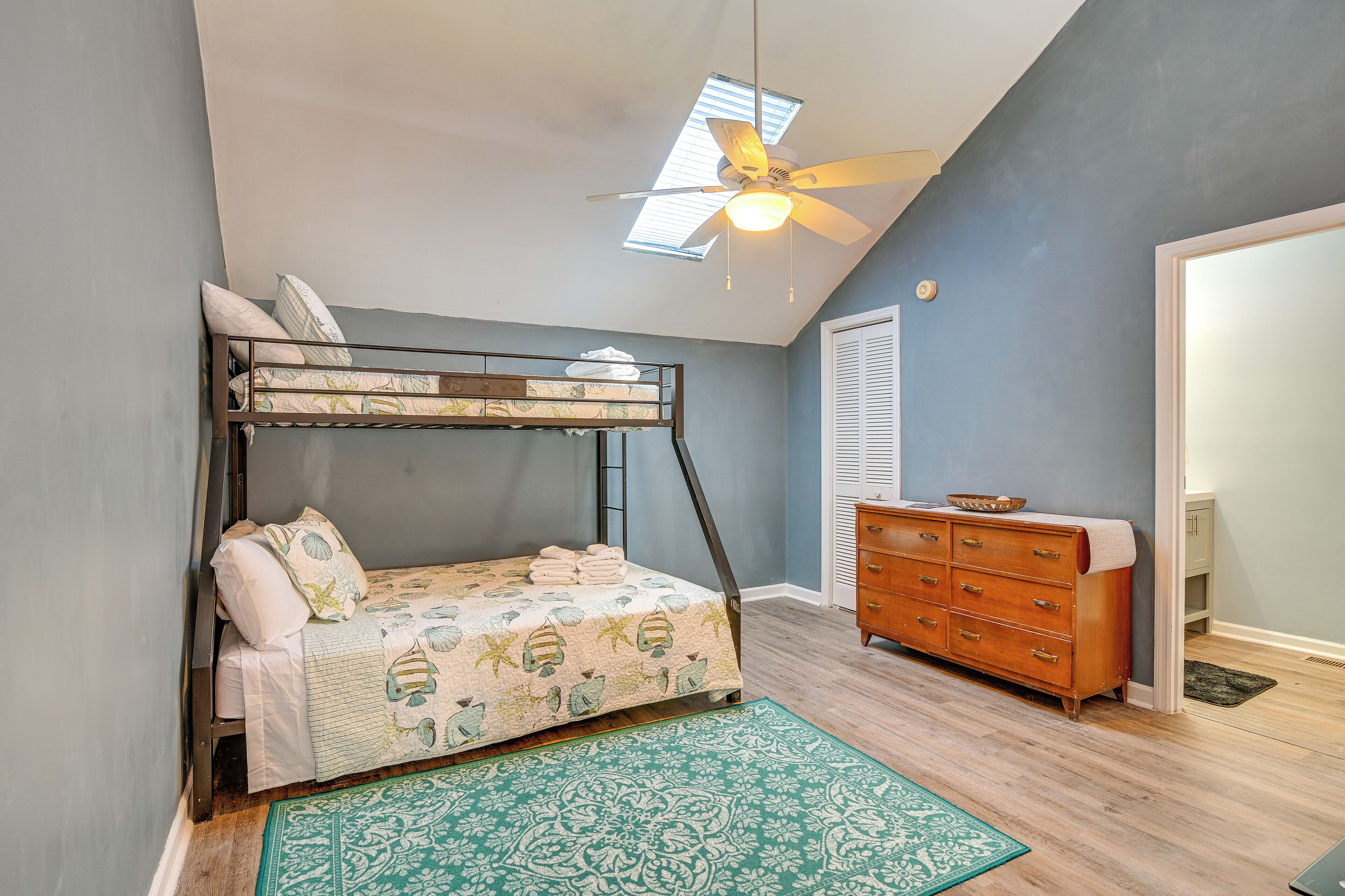 A bunk bed with a patterned bedspread, a wooden dresser, and a ceiling fan in a room with light blue walls and a skylight.