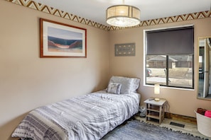 A bedroom with a bed, a framed picture on the wall, a window with a roller blind, a small table with a lamp, and a basket by the door.