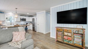 A modern kitchen with white cabinets, a stainless steel refrigerator, and a flat-screen TV mounted on the wall. - < 1 Mi to Beach: Oak Island Retreat w/ Swim Spa! (Oak Island)