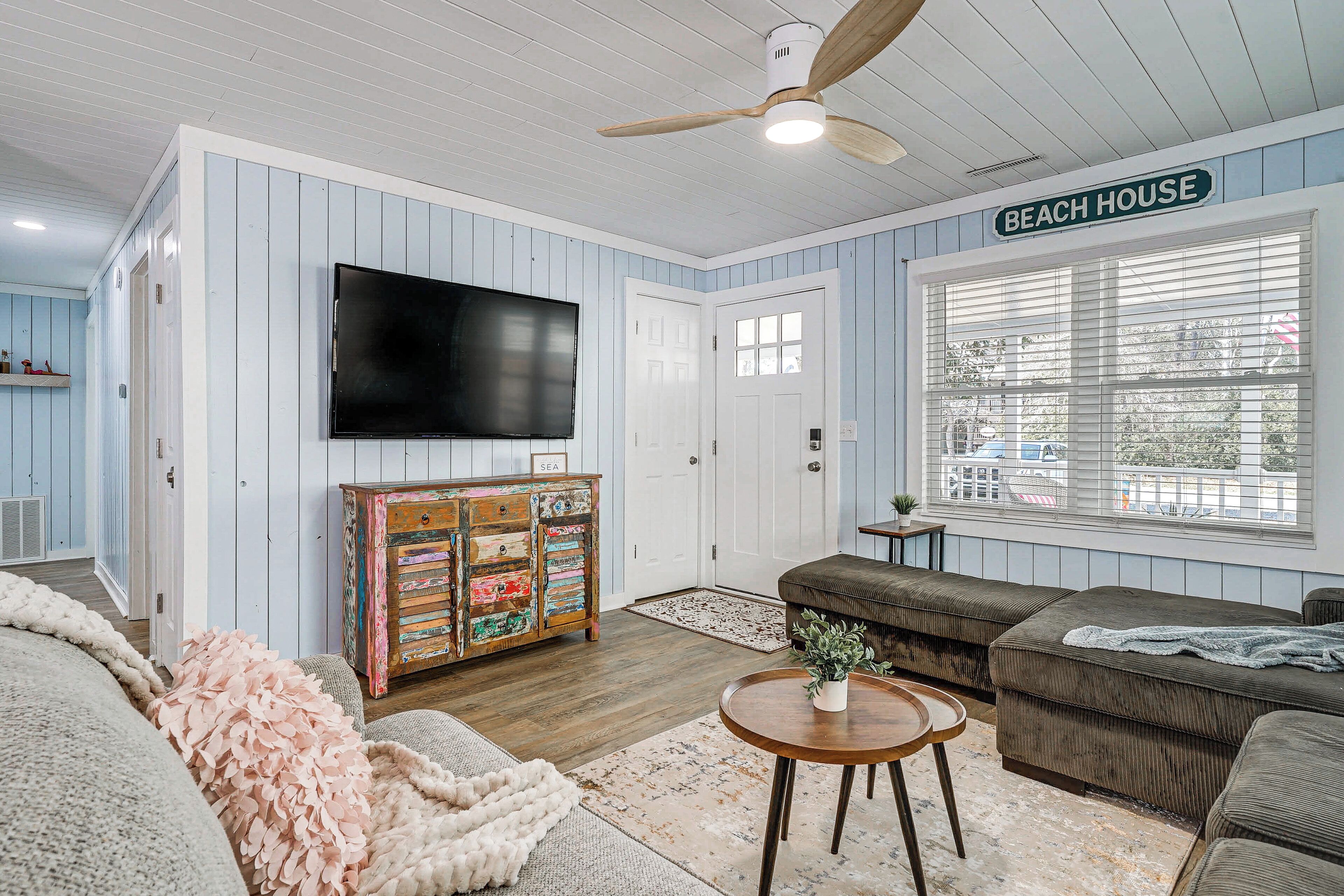 Un salon décoré sur le thème de la plage, avec un téléviseur, un meuble en bois et une grande fenêtre.
