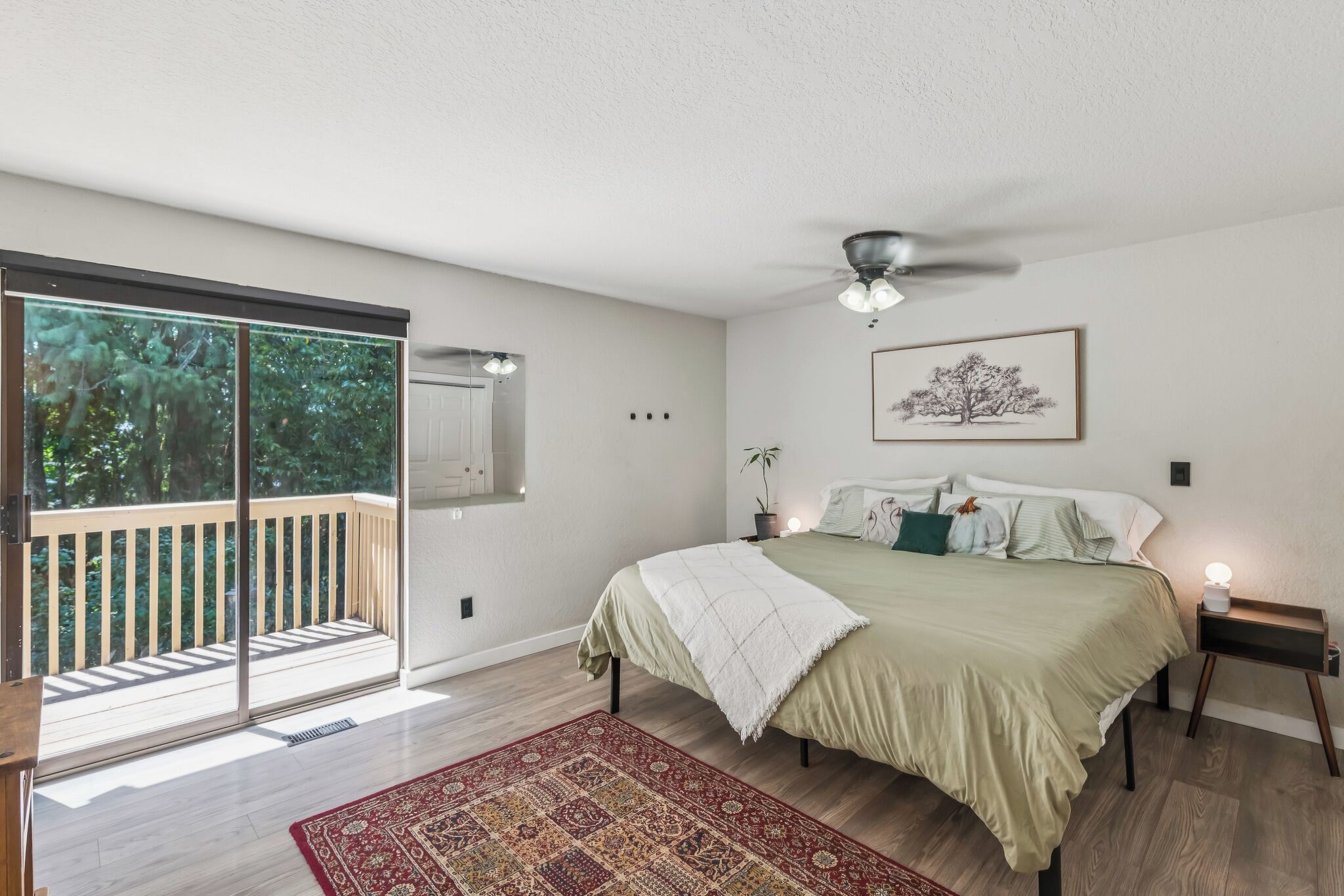 A bedroom with a bed, a ceiling fan, a nightstand, a window with a view of trees, and a red patterned rug.