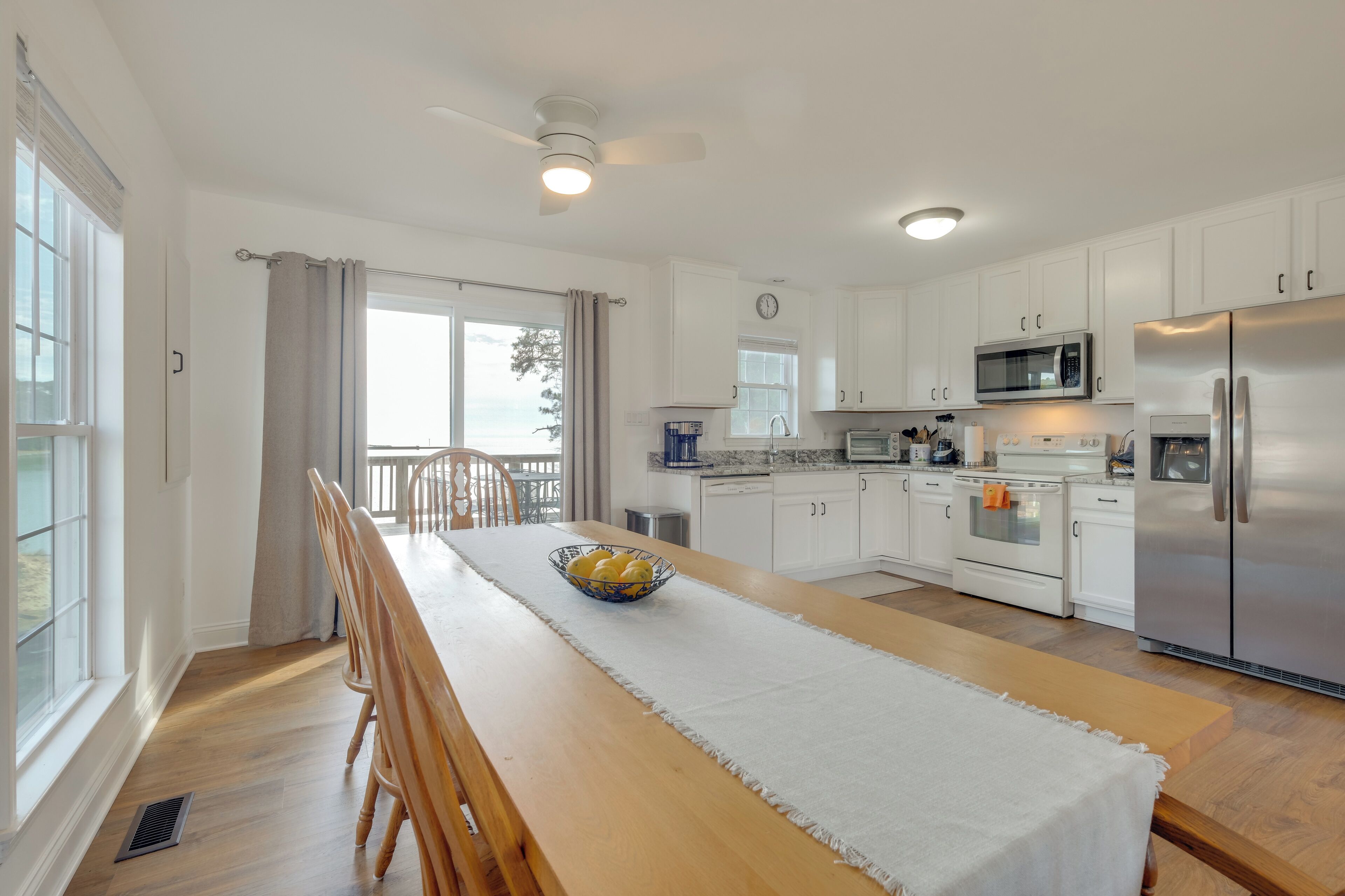 A modern kitchen with white cabinets, stainless steel appliances, and a wooden dining table with a bowl of fruit.