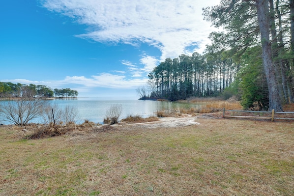 A lakeside view with a wooden fence, grassy area, and tall trees. - Waterfront Gem w/ Deck & Views on Chesapeake Bay! (Reedville)