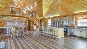 A spacious loft-style interior with a high ceiling, wooden floors, a central island with bar stools, a kitchen area, and a staircase leading to an upper level. - Steps to Private Beach! Family Home on Lake James (Morganton)