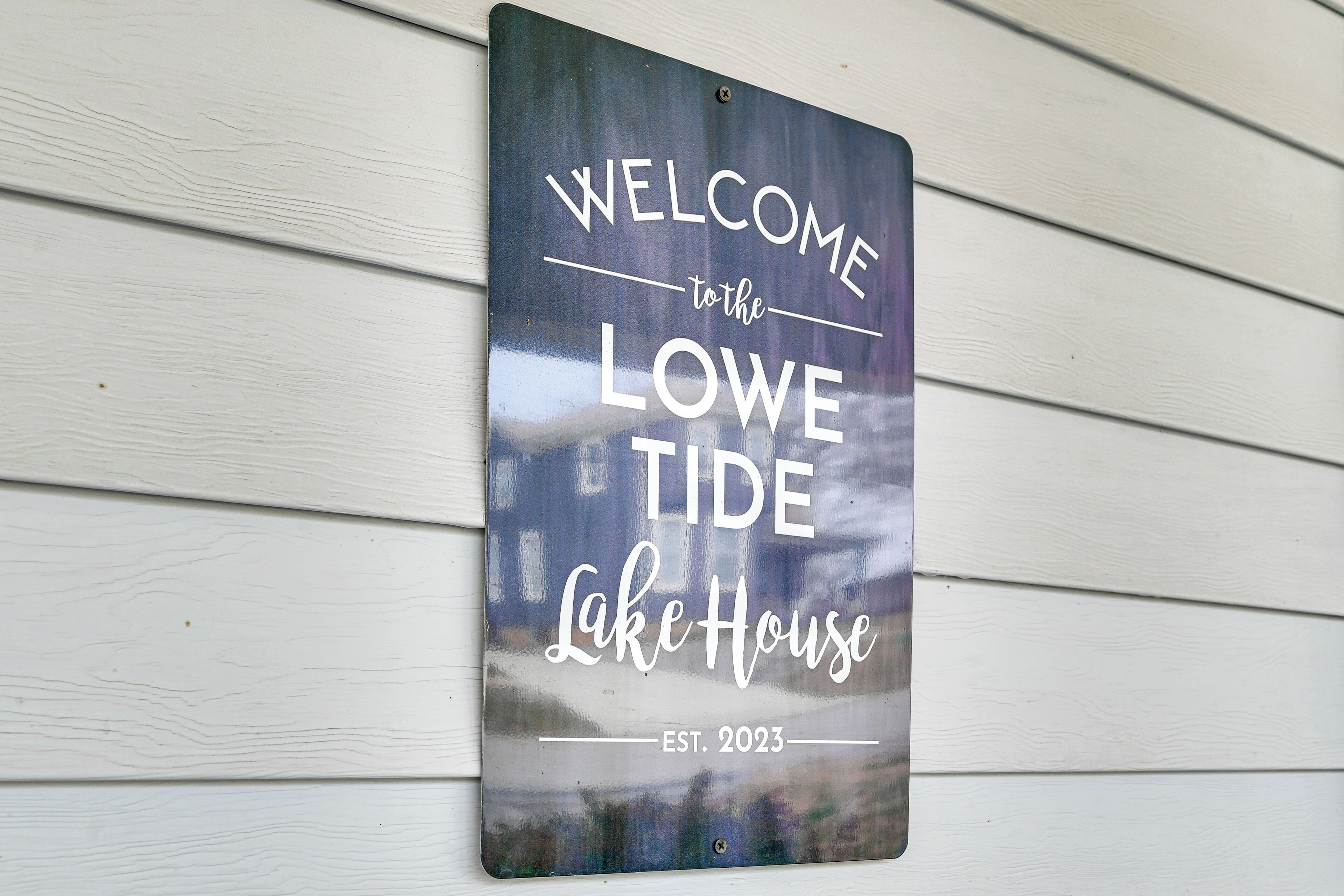A welcome sign for the Lowe Tide Lake House, established in 2023, mounted on a white wooden wall.